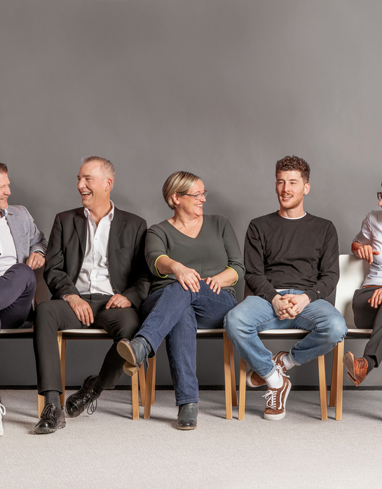 Mitarbeitende des Kantons Solothurn präsentieren sich authentisch, während sie in einer lockeren und freundlichen Atmosphäre auf Stühlen in einer Reihe sitzen und miteinander interagieren.